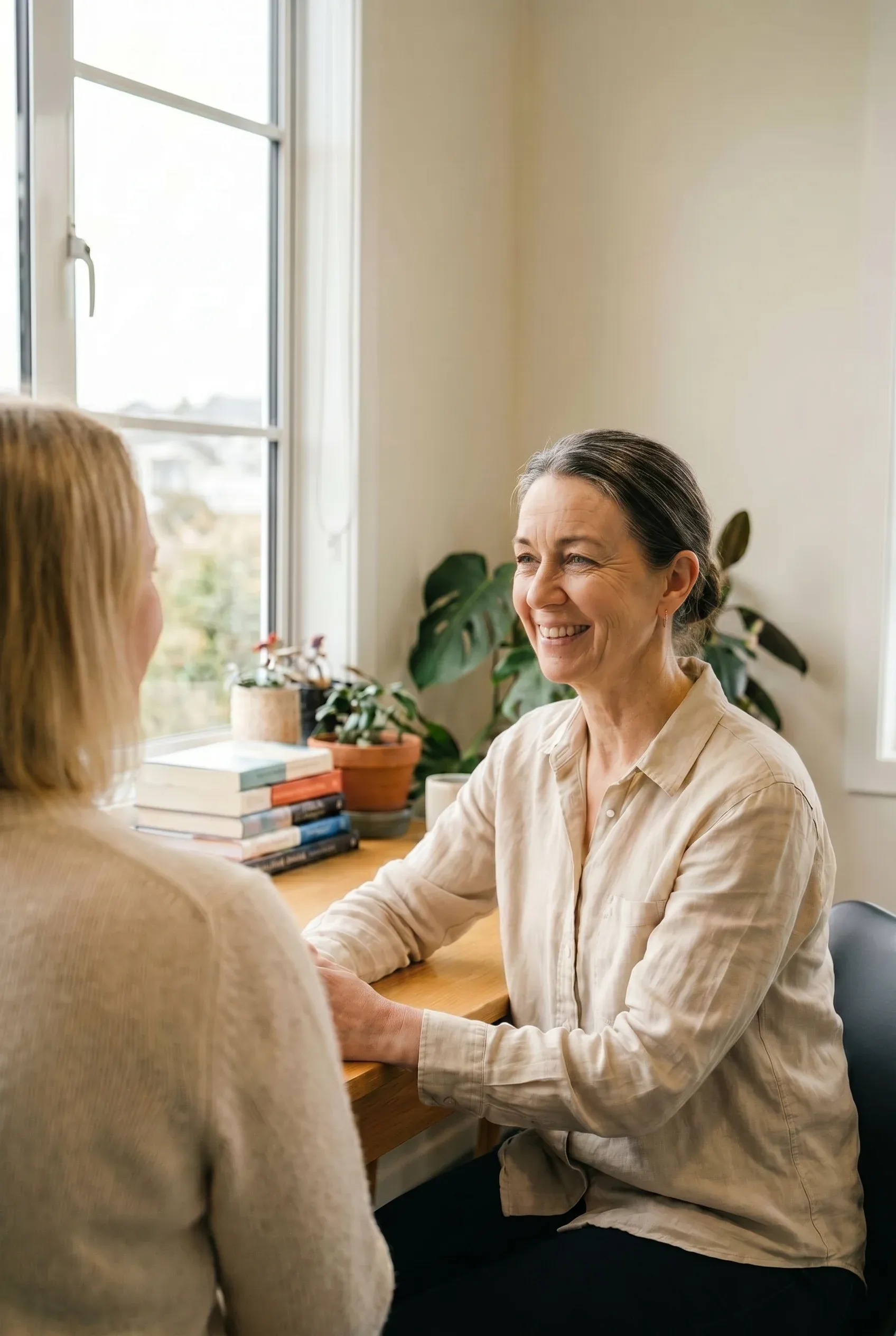 A speech-language therapist in warm conversation with a patient by a sunlit Wellington window